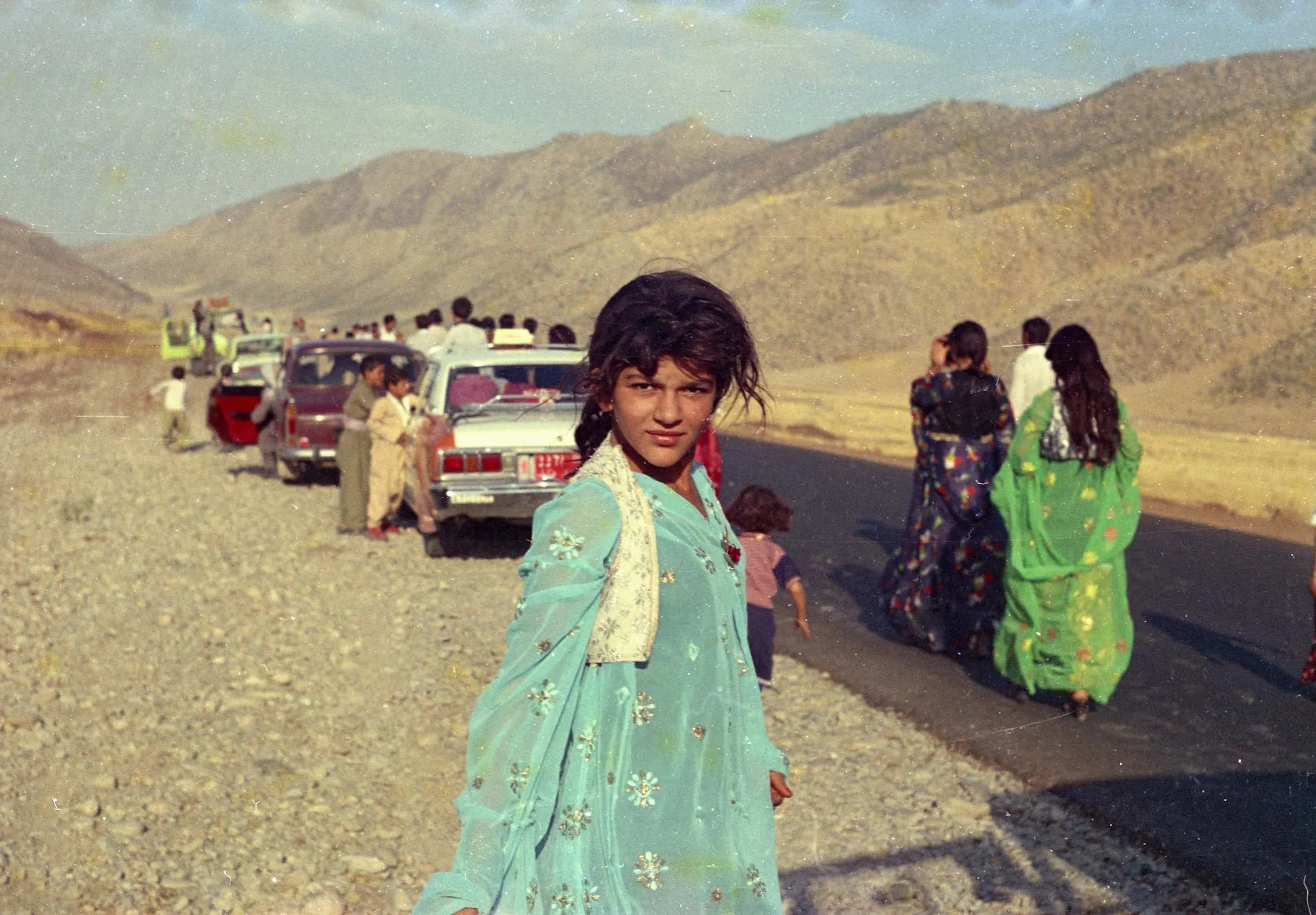 A girl in a pale blue dress stands on a mountain road; behind her a line of parked cars and women in bright dresses.
