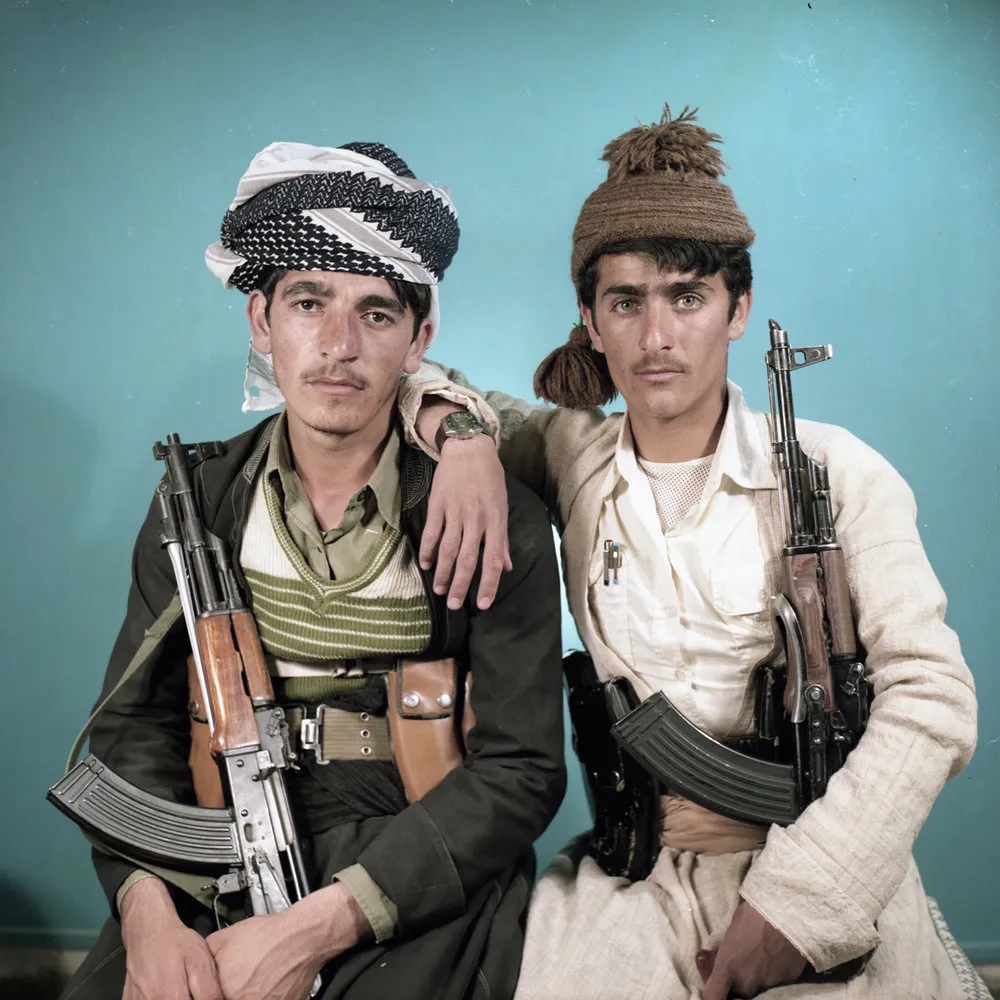 Studio portrait of two young men in Kurdish dress, each holding a rifle, before a turquoise backdrop.