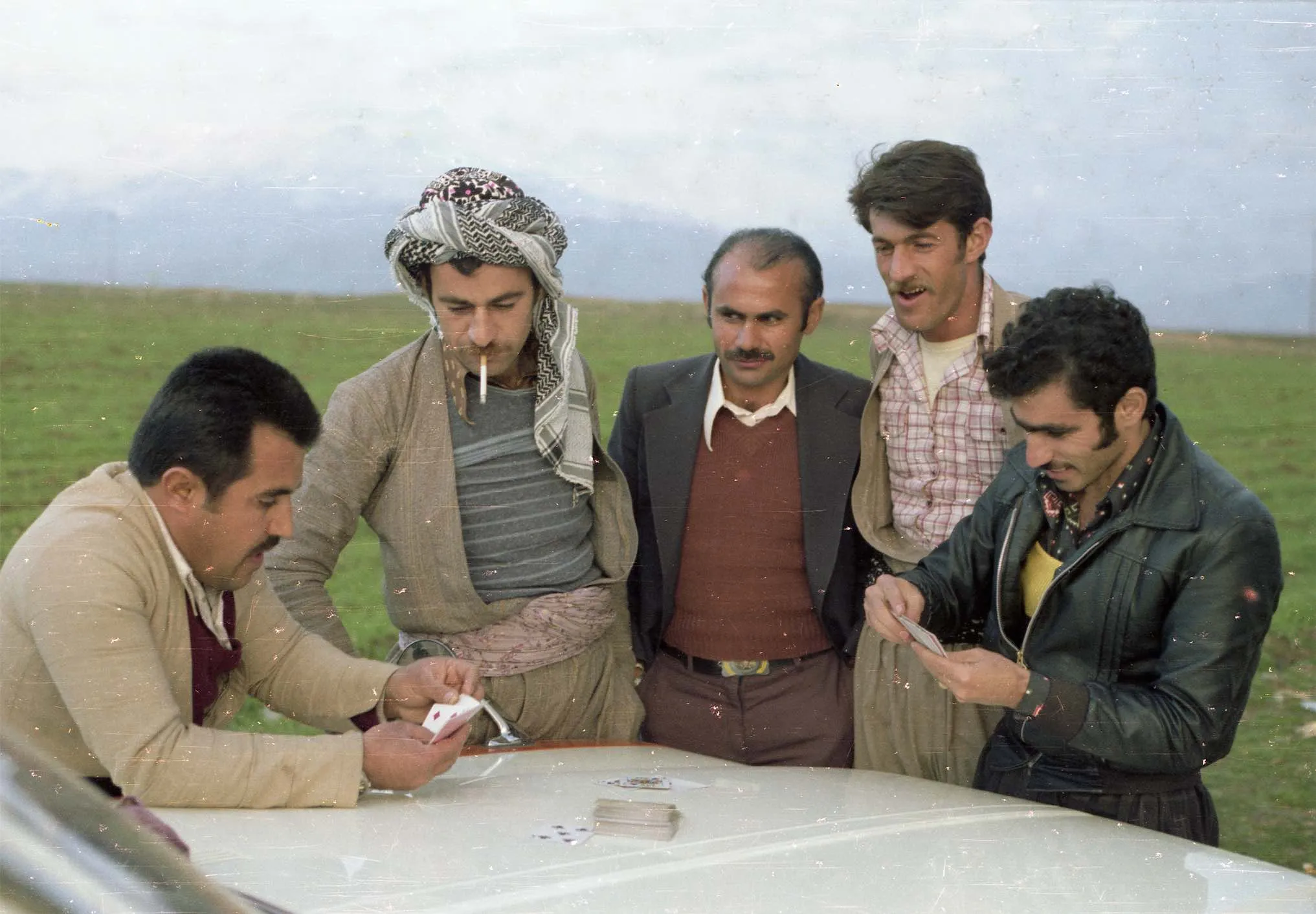 Five men lean over a map spread on the bonnet of a car in open green country.